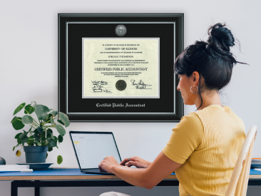 Framed Certified Public Accountant diploma from the University of Illinois, with black matting and silver trim, hanging on a wall next to a woman working on a laptop.