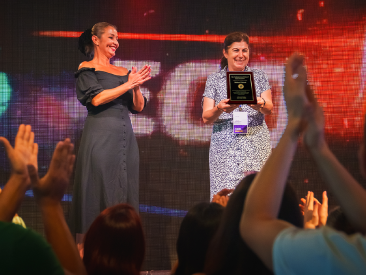 Two women on stage at an awards event; one claps while the other holds a plaque award, with a cheering audience in the foreground.