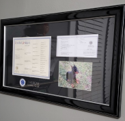 Custom black gloss frame featuring a three-piece American Kennel Club display, including a certified pedigree certificate, registration paper, and a photo of a black and tan dog lying on grass. The AKC logo is displayed on the black matting.