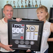 Two people proudly holding a large custom Inc. 500 award plaque featuring magazine covers, company stats, and logos, with trophies visible in the background.