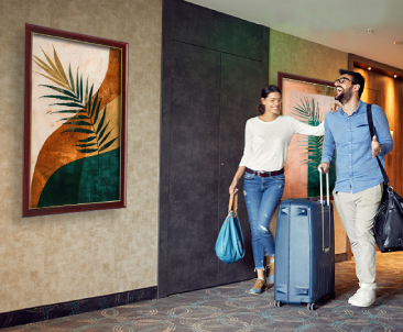 Framed abstract art prints with green palm leaves and warm-toned backgrounds, hanging in a hallway as a smiling couple walks by with luggage.