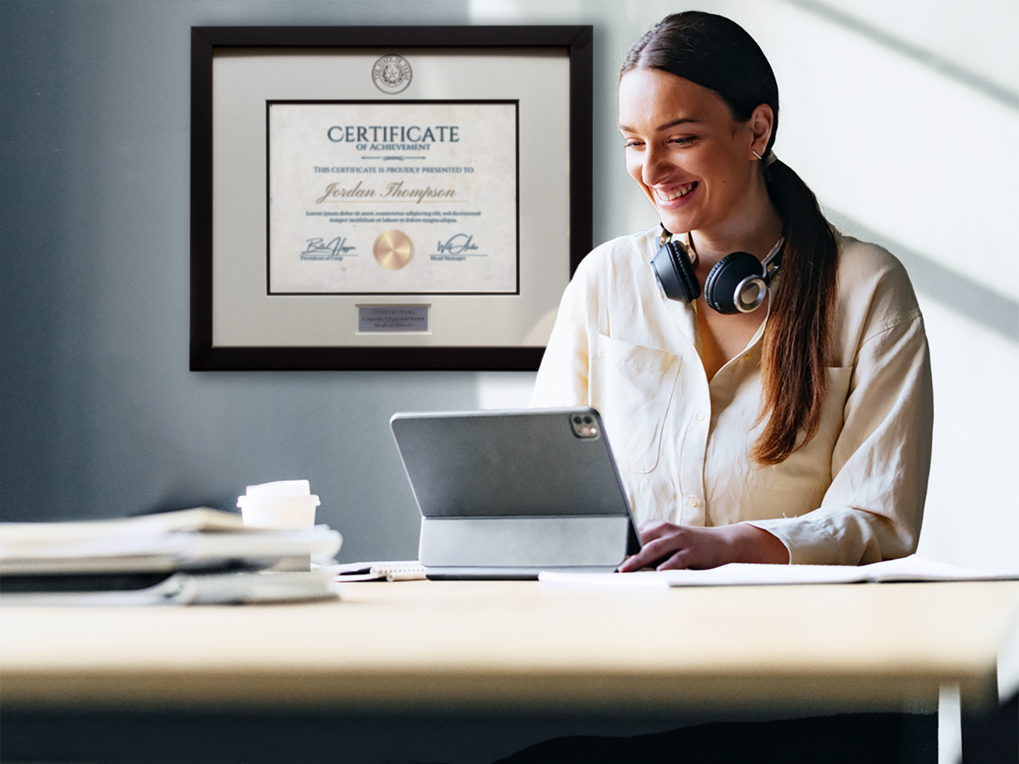 Smiling woman working on a tablet at a desk, with a framed Certificate of Achievement displayed on the wall behind her.